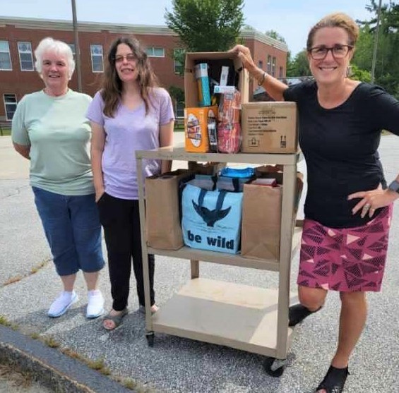 Three ladies standing near a rolling cart filled with school supplies.