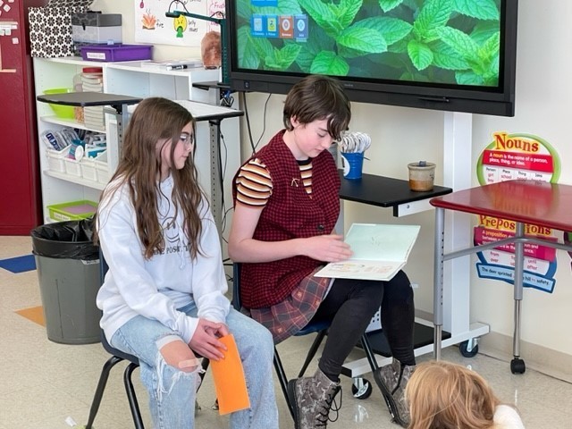 Two students reading  "All Are Welcome" to students sitting on a rug out of the frame