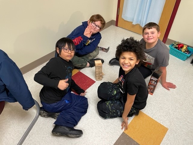 A group of four third grade boys play Jenga on the floor