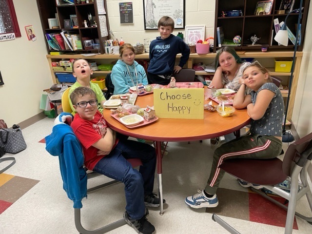 a group of fifth graders sit at a table together for lunch with a choose happy sign