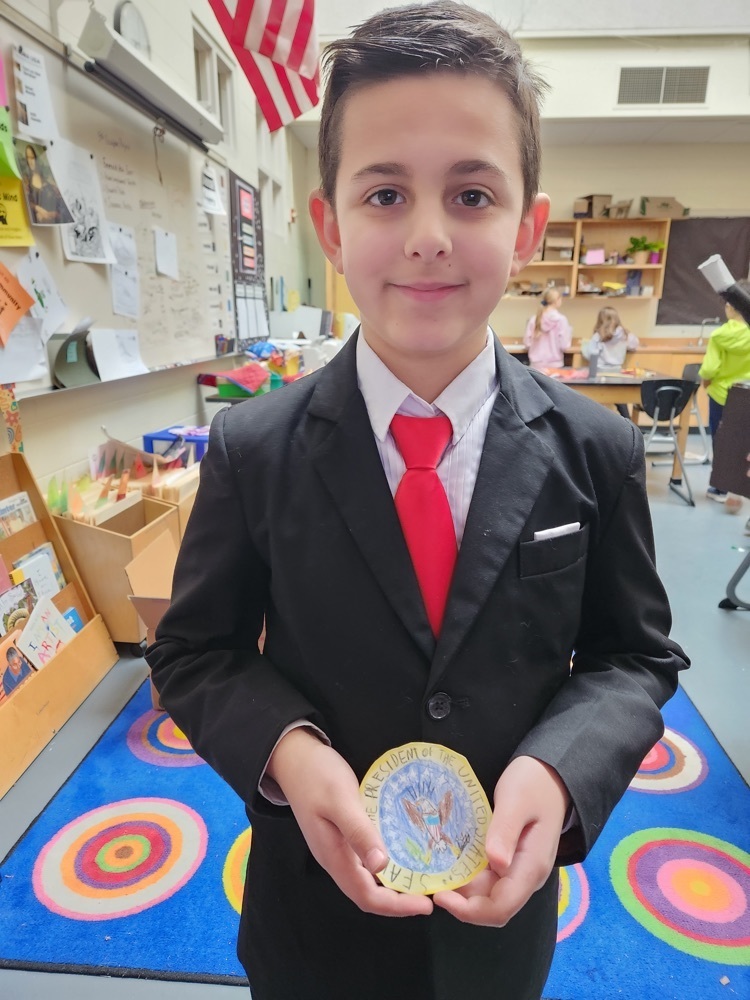 5th grader wearing a black suit with a white shirt and red tie holds a Presidential Seal he created